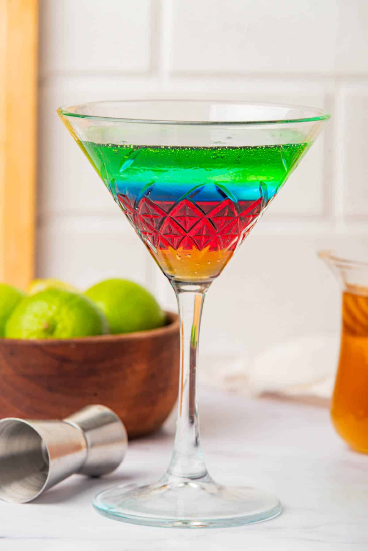Martini glass with liquid in the colors of the Olympic Rings with a jigger on side, limes, bottles, and honey in background.