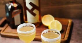 Two cocktails with sugared rims with bottles of liqueur and a lemon on cutting board in background for Pinterest graphic.