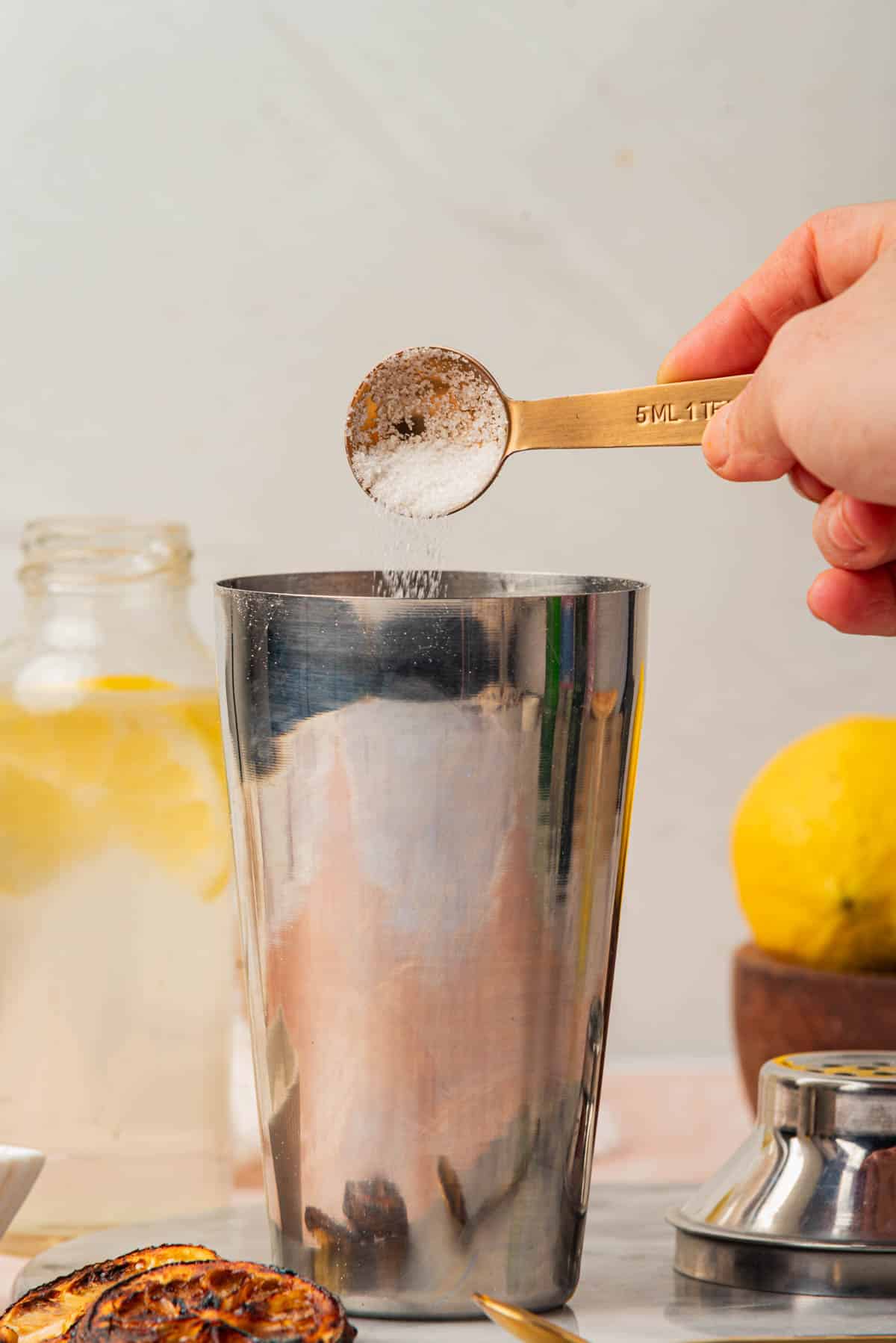 Pouring sugar into cocktail shaker with and lemon slices, bowl of lemons, and spoon in background.