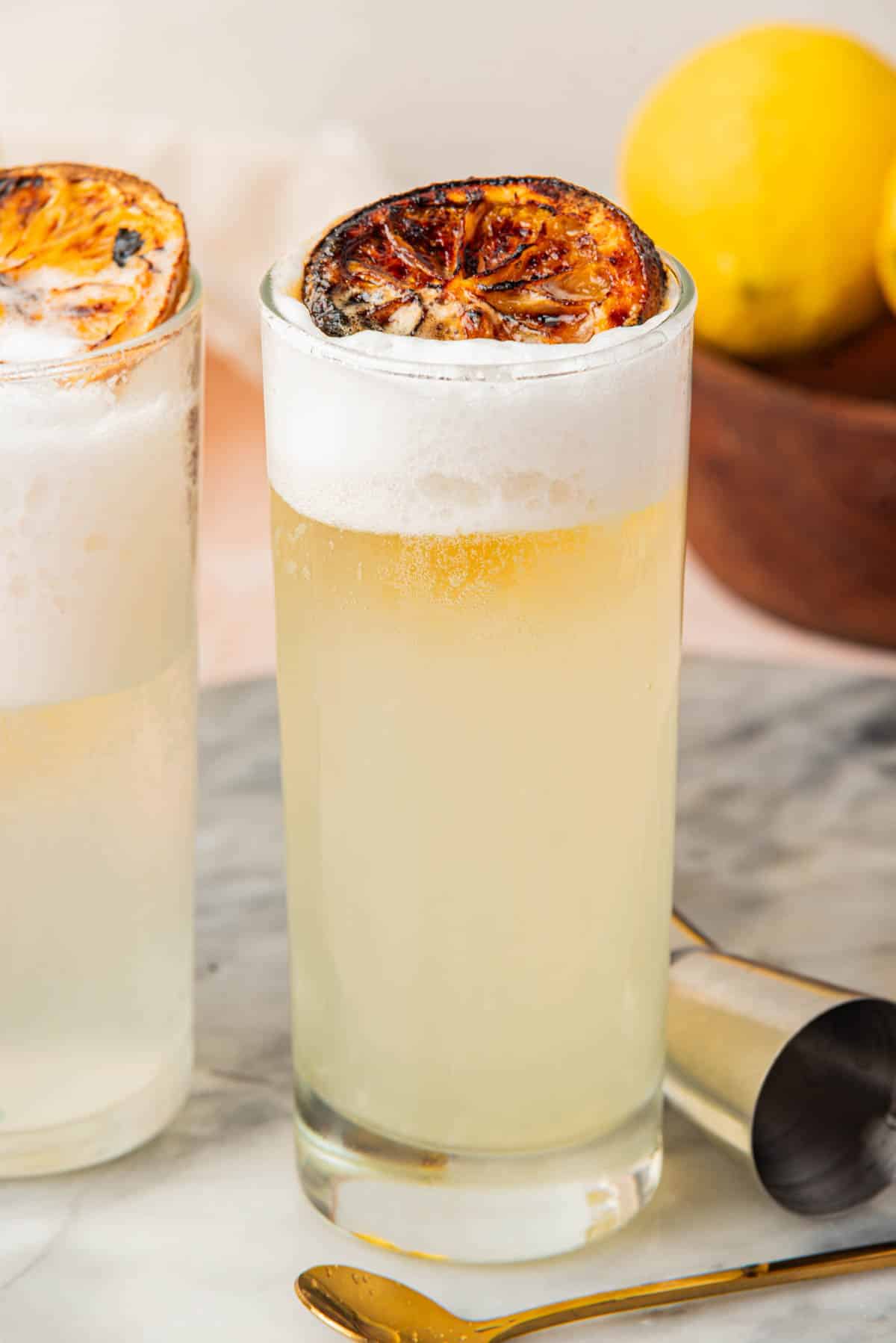 Cocktails with foam and lemon slice on top with bowl of lemons, silver cocktail shaker, jigger, spoon, and bowl with small spoon in background.