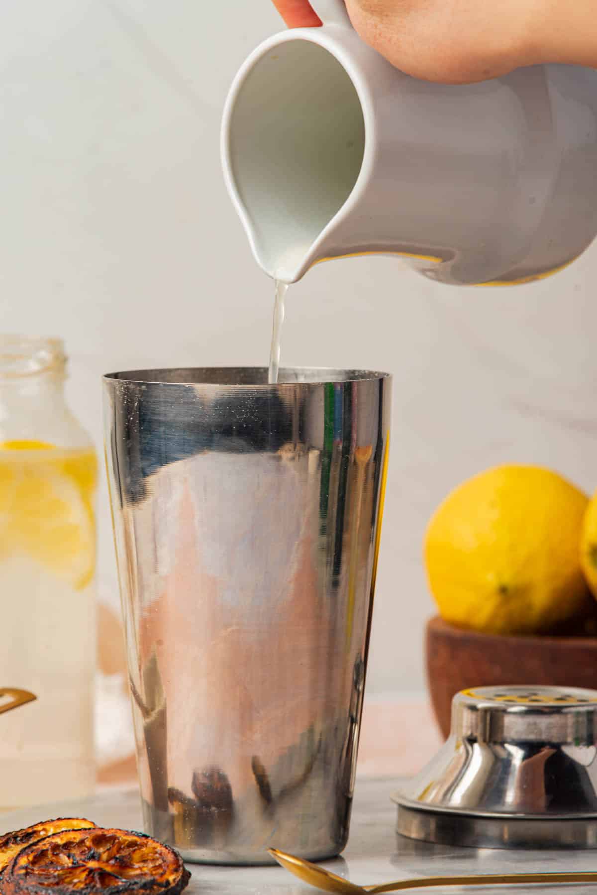 Pouring lemon juice into cocktail shaker with and lemon slices, bowl of lemons, and spoon in background.