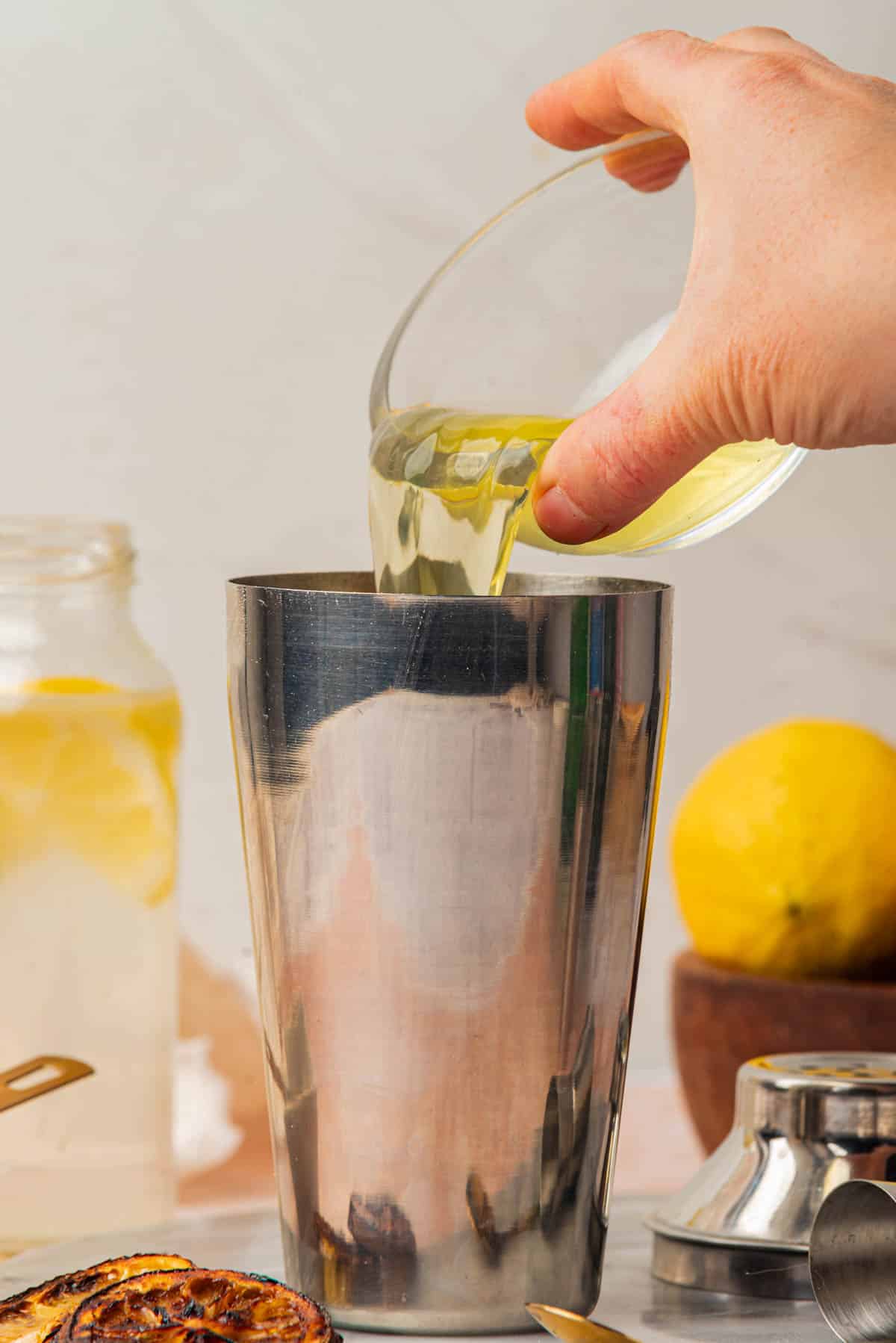 Pouring egg whites into glass with and lemon slices, bowl of lemons, jigger, and spoon in background.