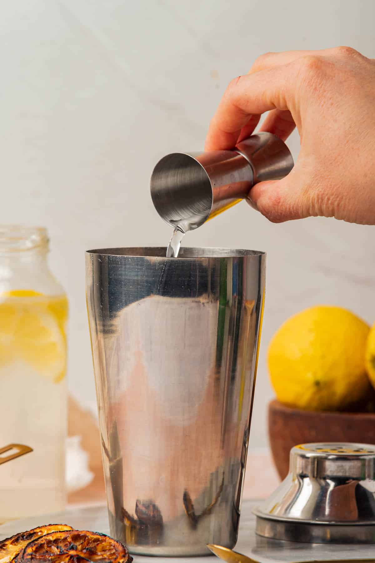 Pouring gin into cocktail shaker with and lemon slices, bowl of lemons, and spoon in background.