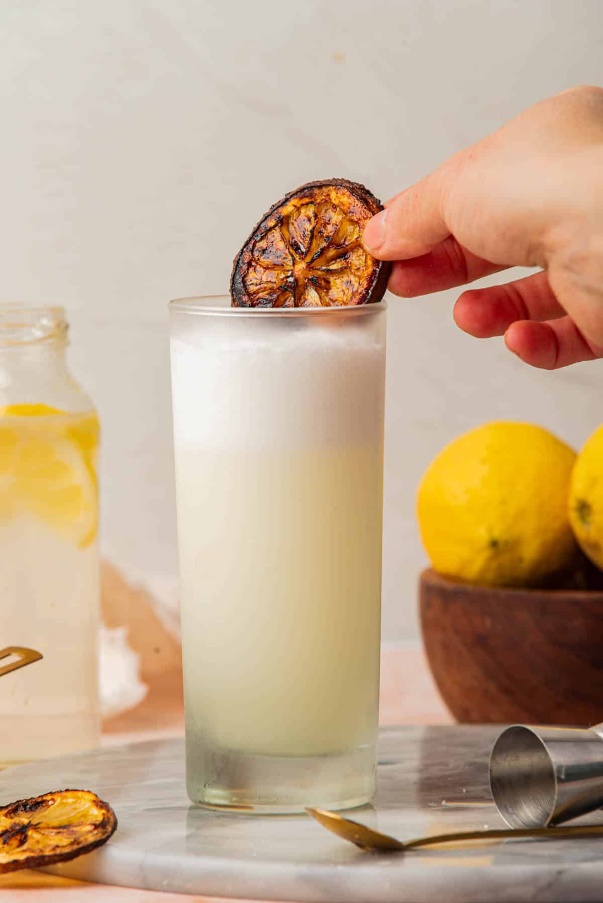 Cocktails with foam and lemon slice on top with bowl of lemons, silver cocktail shaker, jigger, and spoon.
