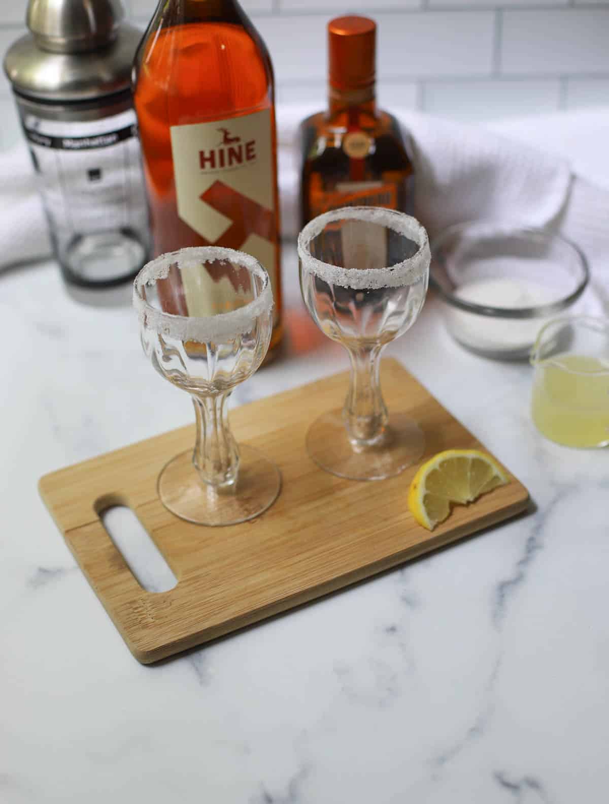 Two glasses rimmed with sugar on cutting board with lemon with bottles and shaker in background.