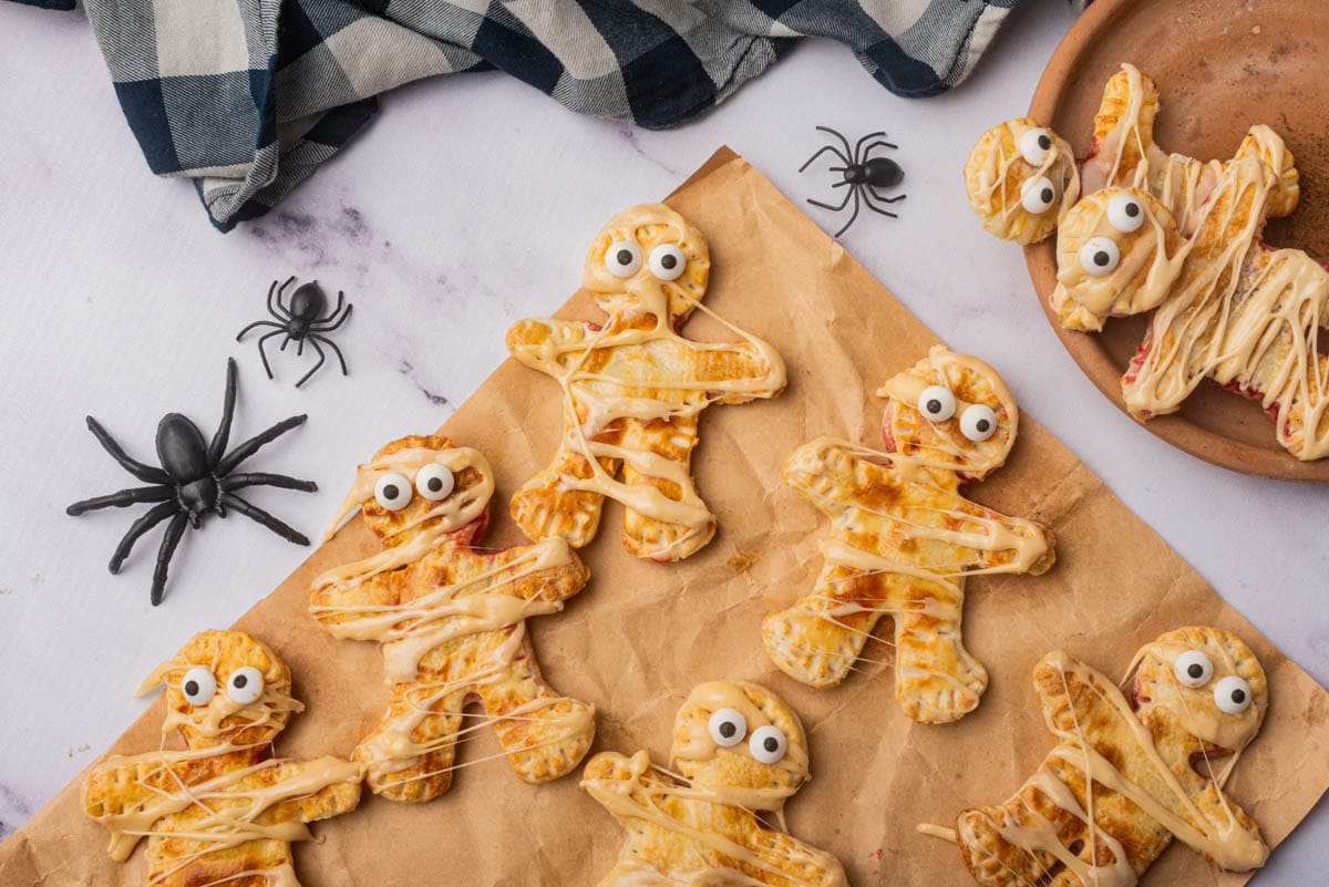 Mummy shaped individual pies with icing drizzle on paper on table with spiders and black and white napkin.