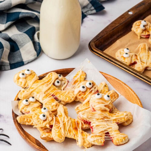 Mummy shaped individual pies with icing drizzle on paper on table with spiders and black and white napkin.