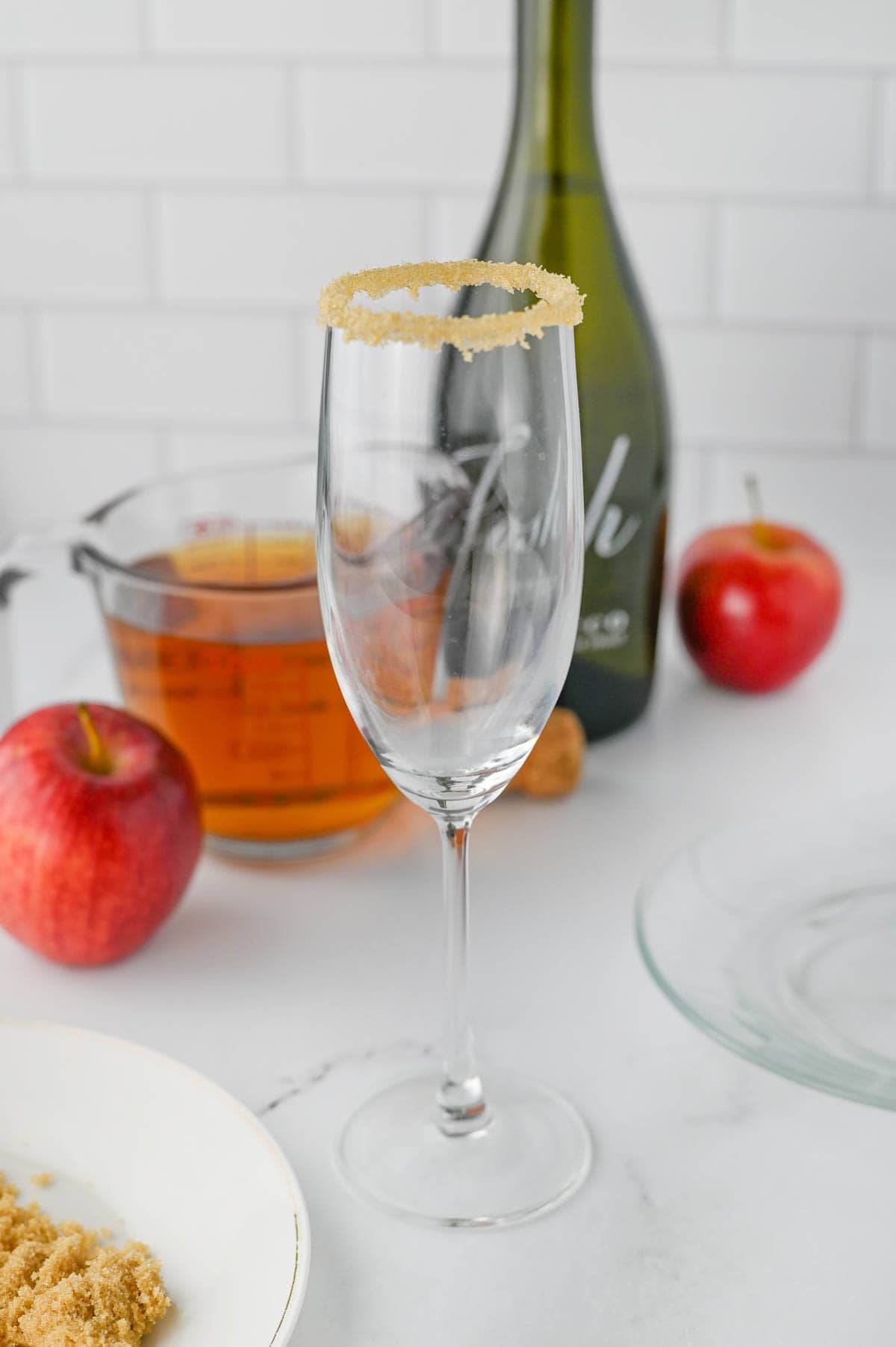 Champagne flute with brown sugar rim and apple cider in glass measuring cup, bottle of Prosecco, and apples in background.