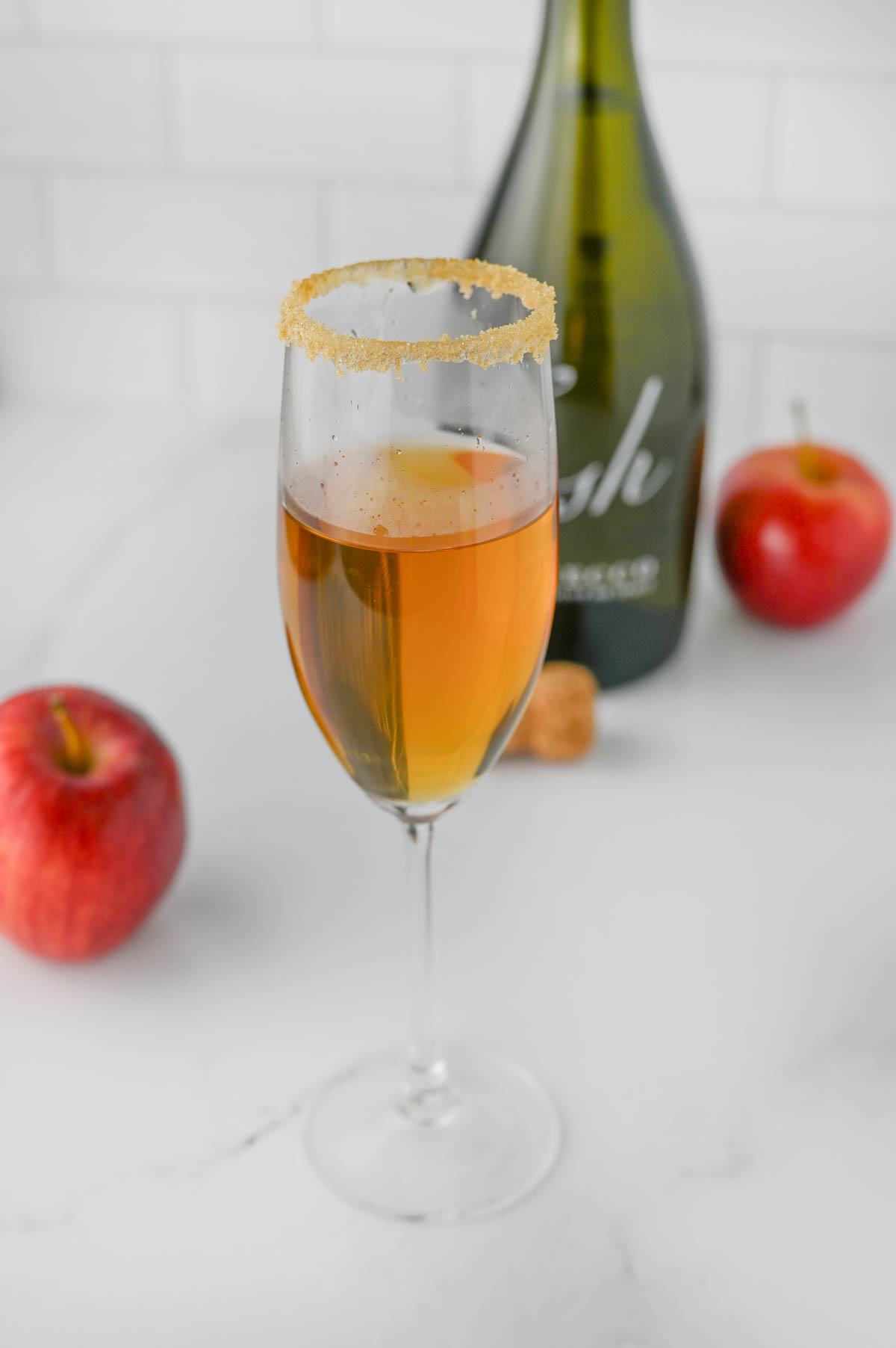 Champagne flute filled halfway with apple cider, apples, and bottle of Prosecco in background.