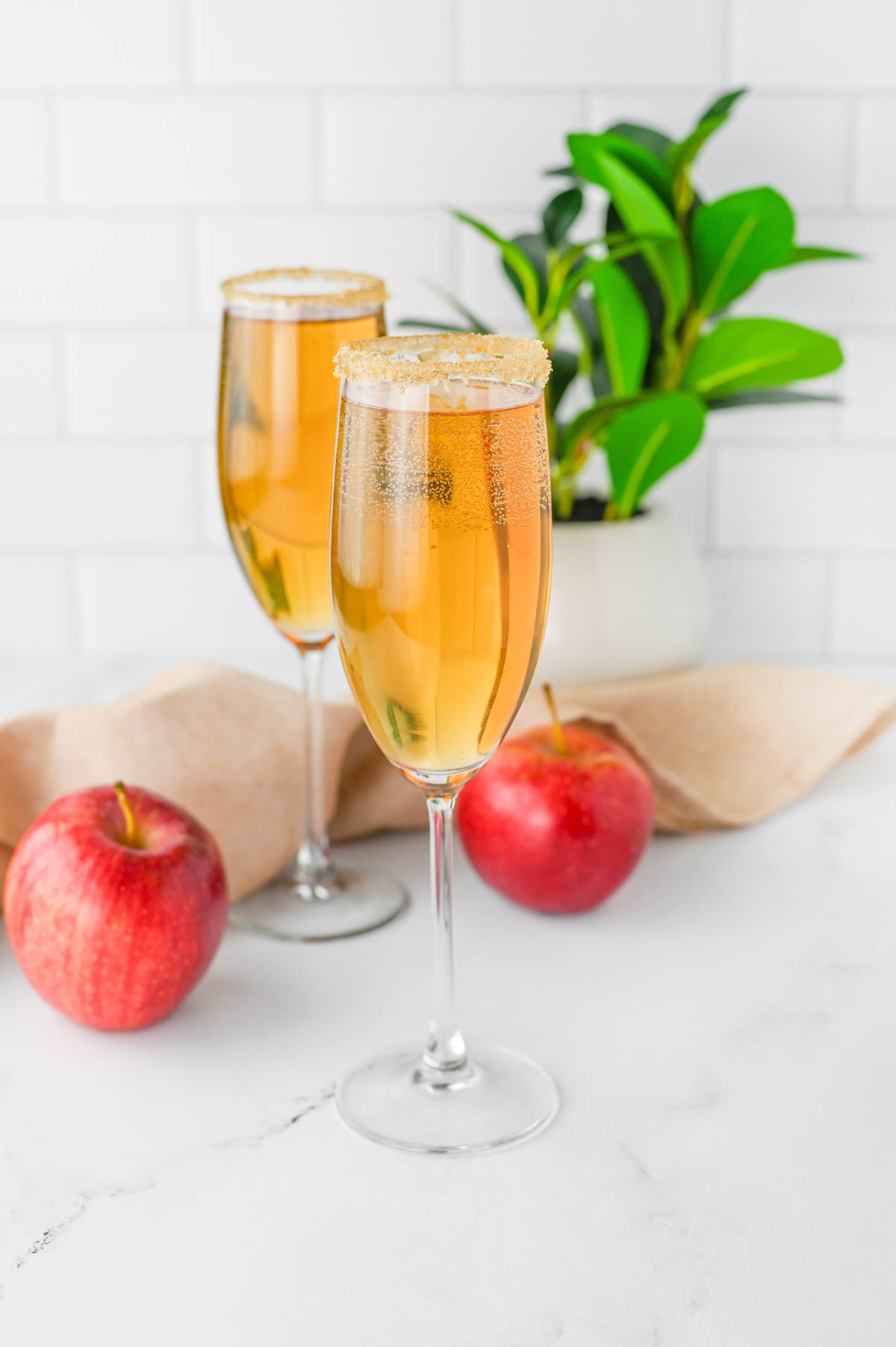 Champagne flutes with apple cider and Prosecco with apples and cloth napkin and plant in background.