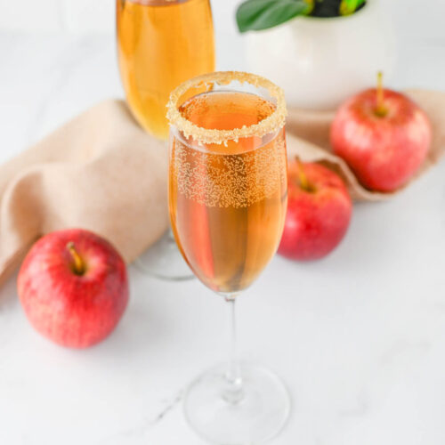 Champagne flutes with apple cider and Prosecco with apples and cloth napkin and plant in background.