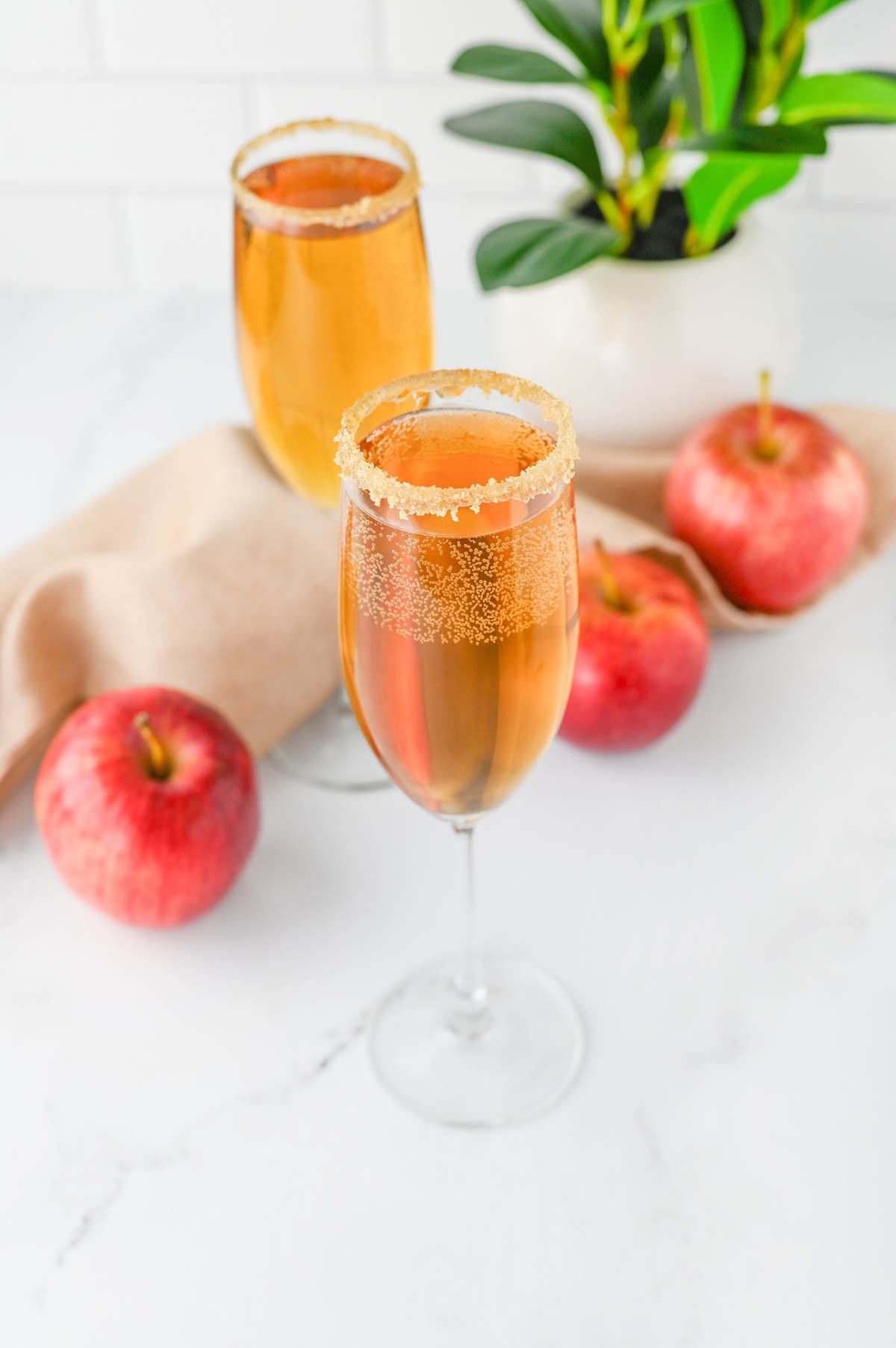 Champagne flutes with apple cider and Prosecco with apples and cloth napkin and plant in background.