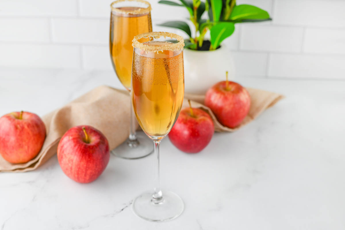 Champagne flutes with apple cider and Prosecco with apples and cloth napkin and plant in background.