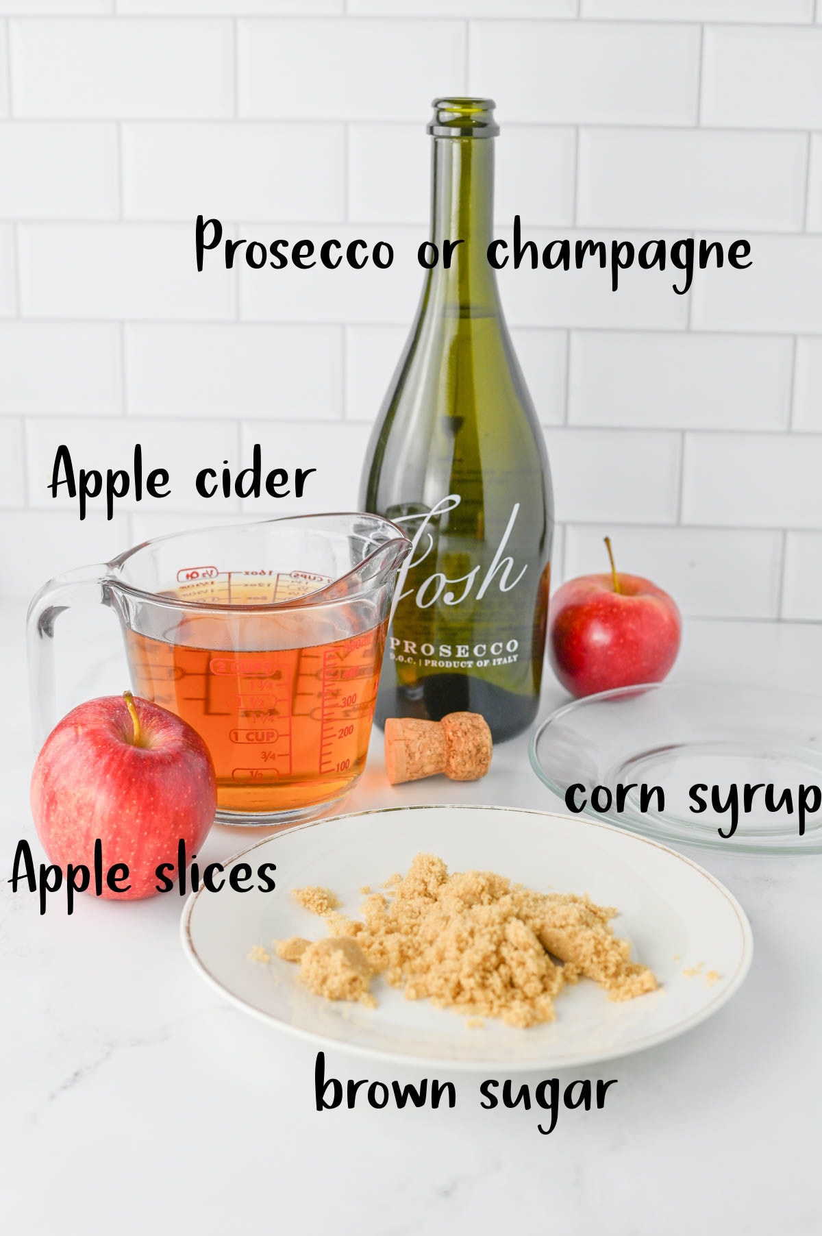 Prosecco with cork in front, apple cider, apples, brown sugar on white plate, and glass plate.