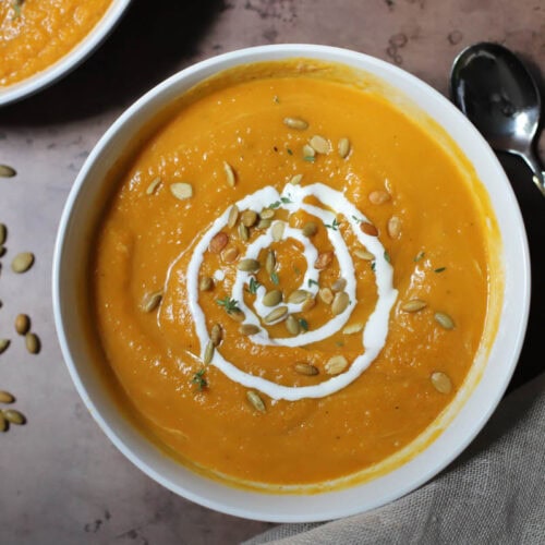 Butternut squash soup topped with sour cream and pepitas in a white bowl on a brown counter.