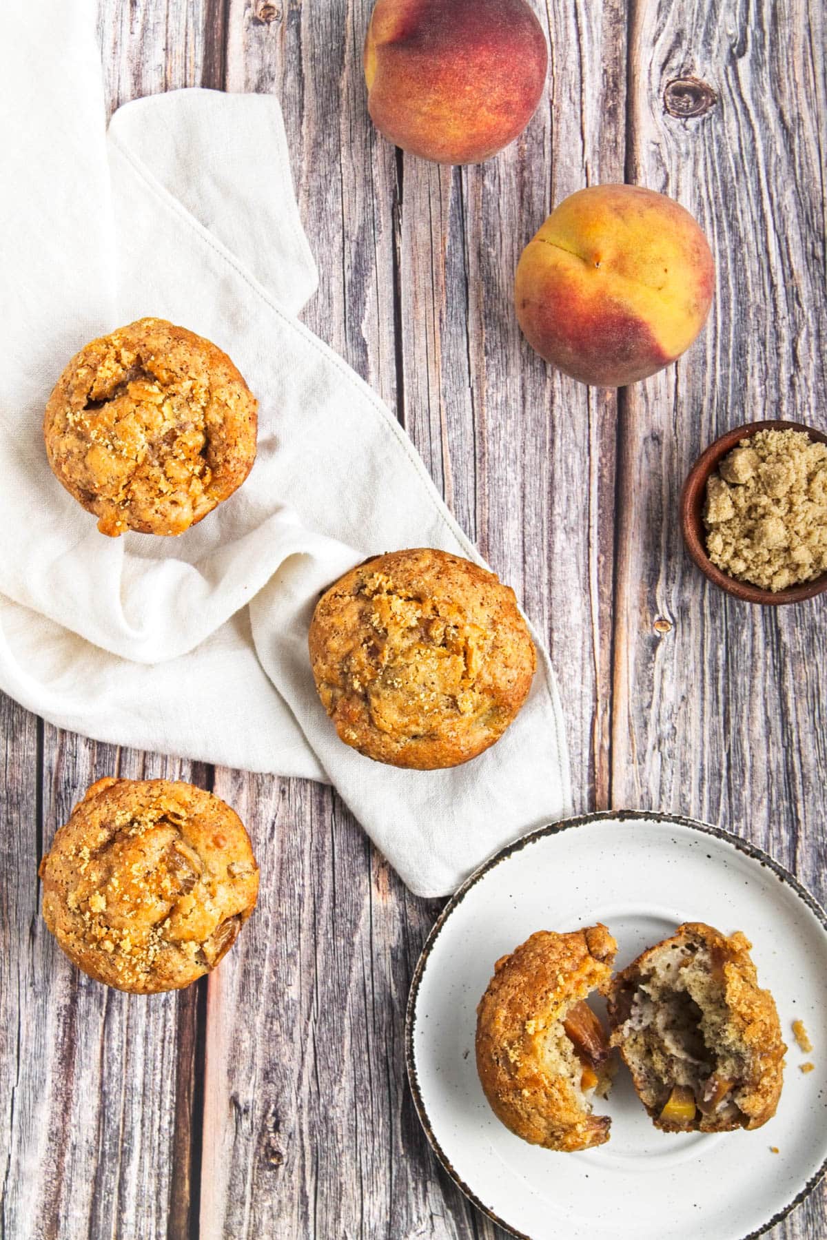 Peaches and muffins on wood table with bowl of brown sugar and one muffin on white plate.