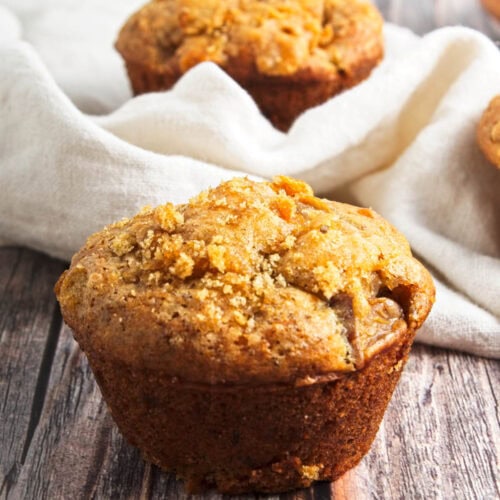 Muffin on wood board with another muffin in napkin in background.