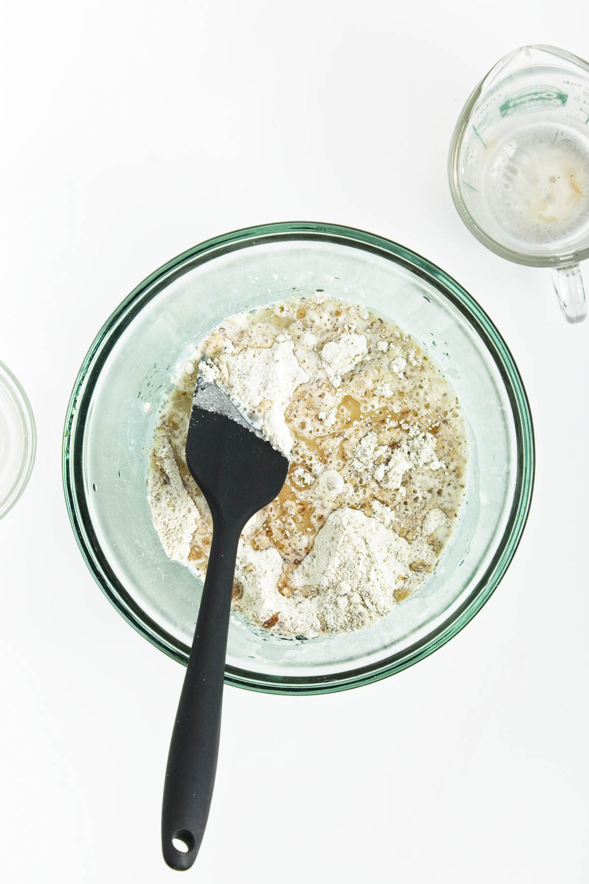 Flour and liquid in glass bowl with glass measuring cup in back.