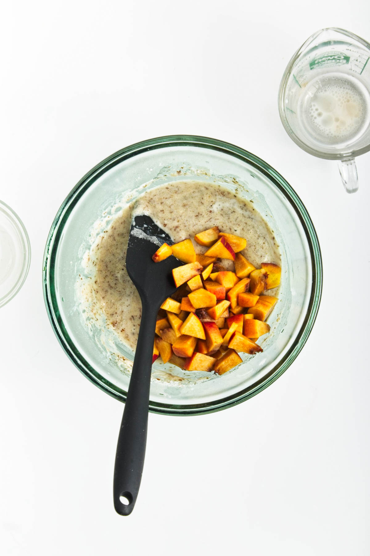 Muffin batter with chopped peaches in glass bowl with glass measuring cup in background.