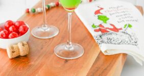 Green liquid in a champagne flute with red cherry in bottom of glass, with Grinch napkin on wood board on Pinterest Pin image.