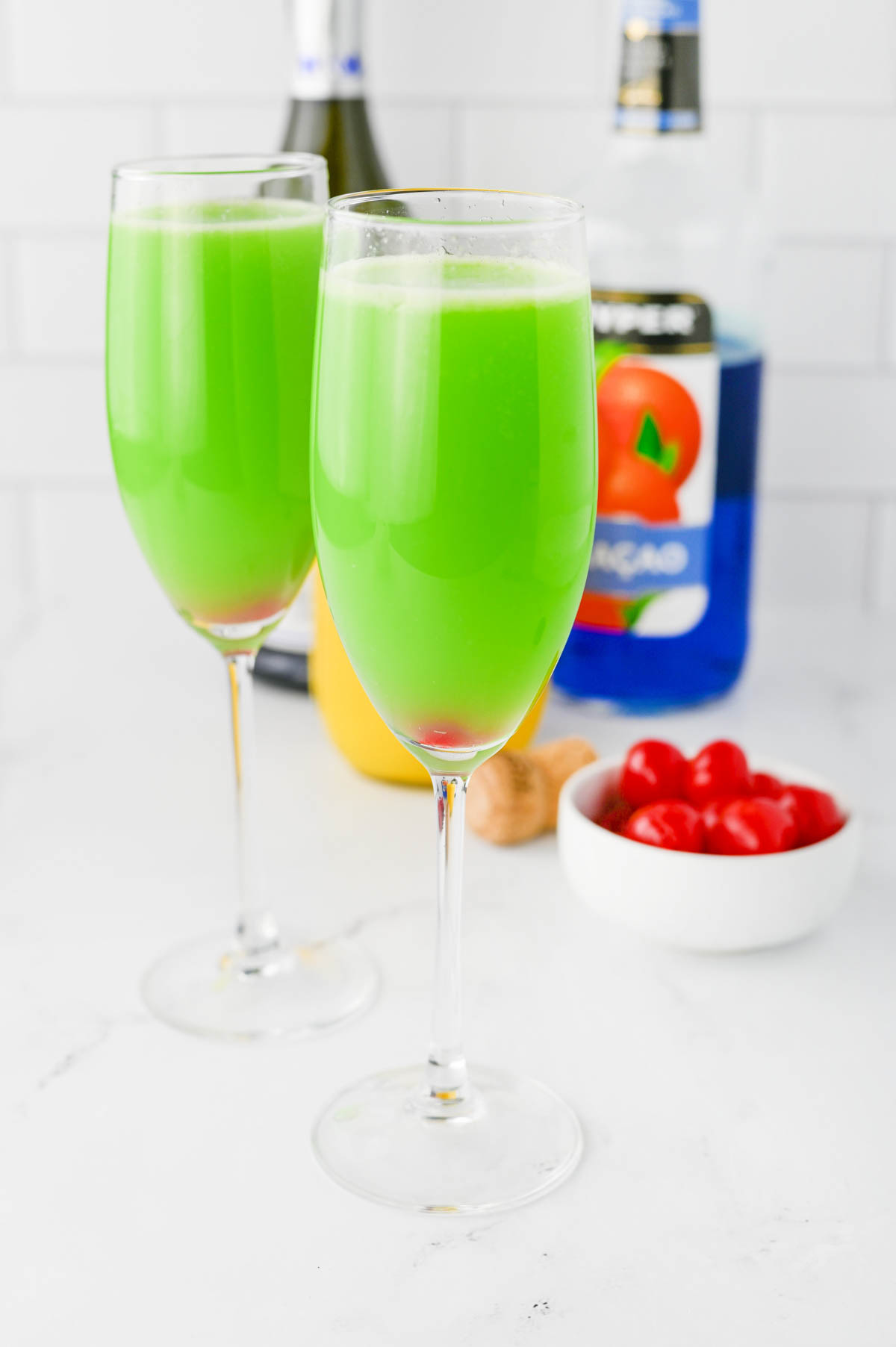 Green liquid in a champagne flute with red cherry in bottom of glass, with Grinch napkin on white counter.