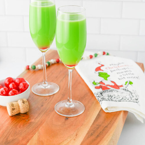 Green liquid in a champagne flute with red cherry in bottom of glass, with Grinch napkin on wood board.