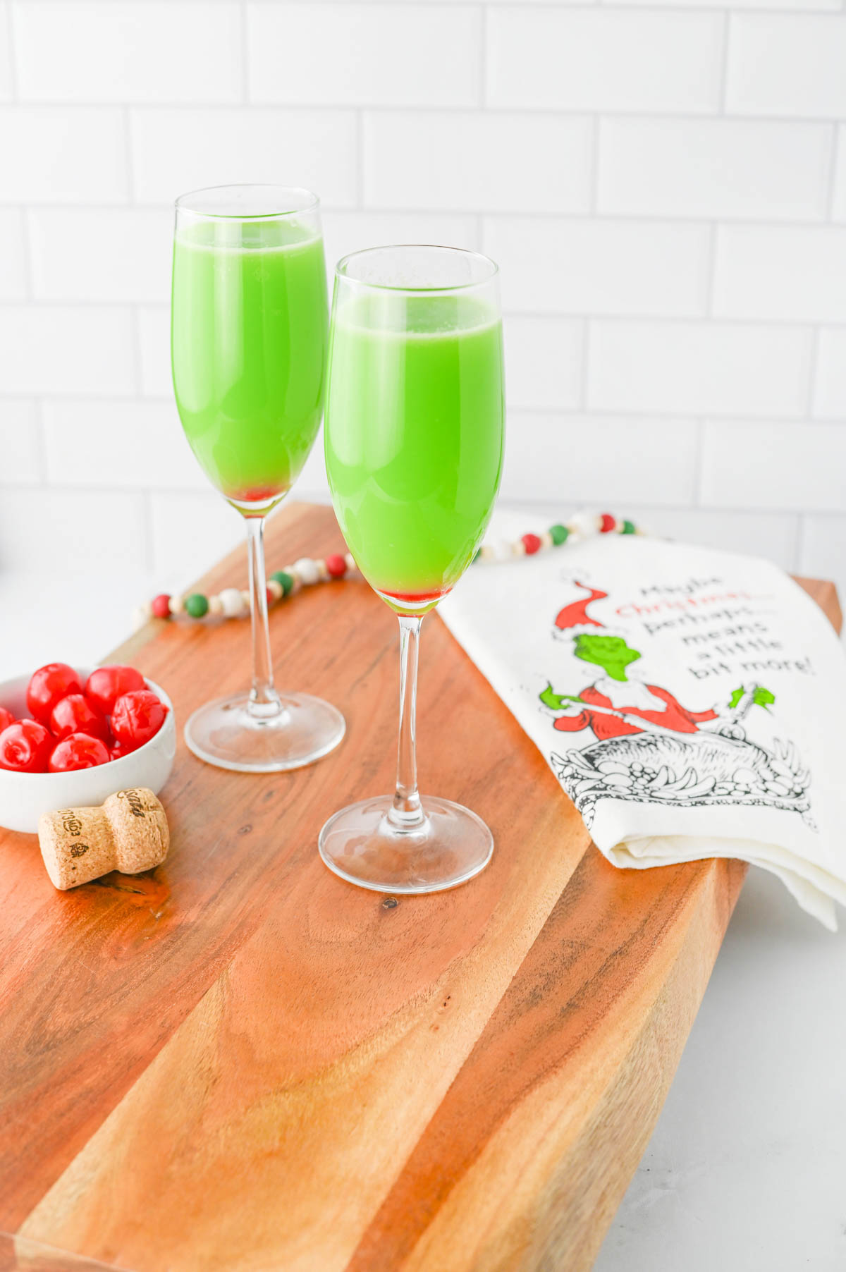 Green drink with red bottom in a champagne flute with bowl of cherries and Grinch napkin on wood board.