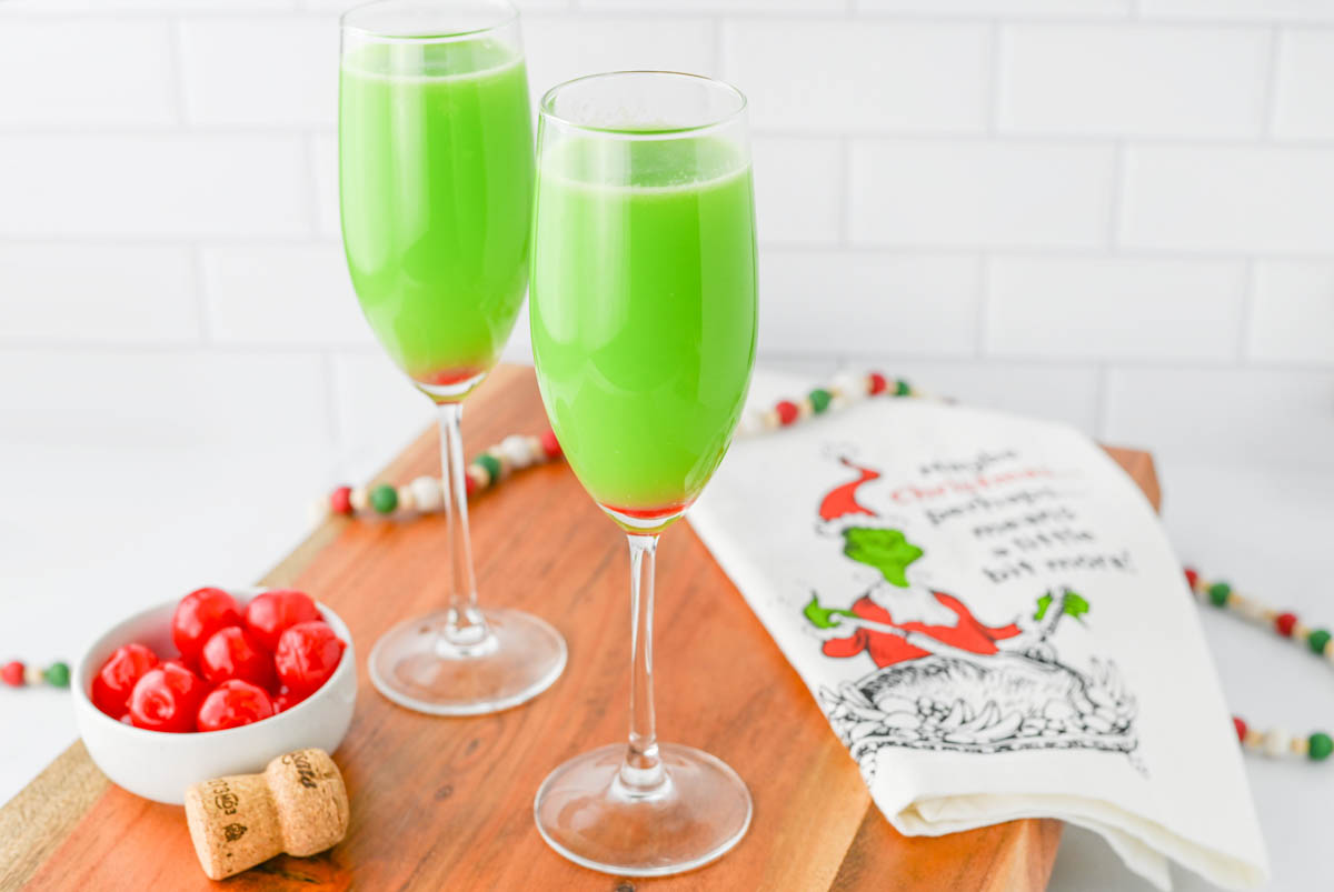 Green liquid in a champagne flute with red cherry in bottom of glass, with Grinch napkin on wood board.