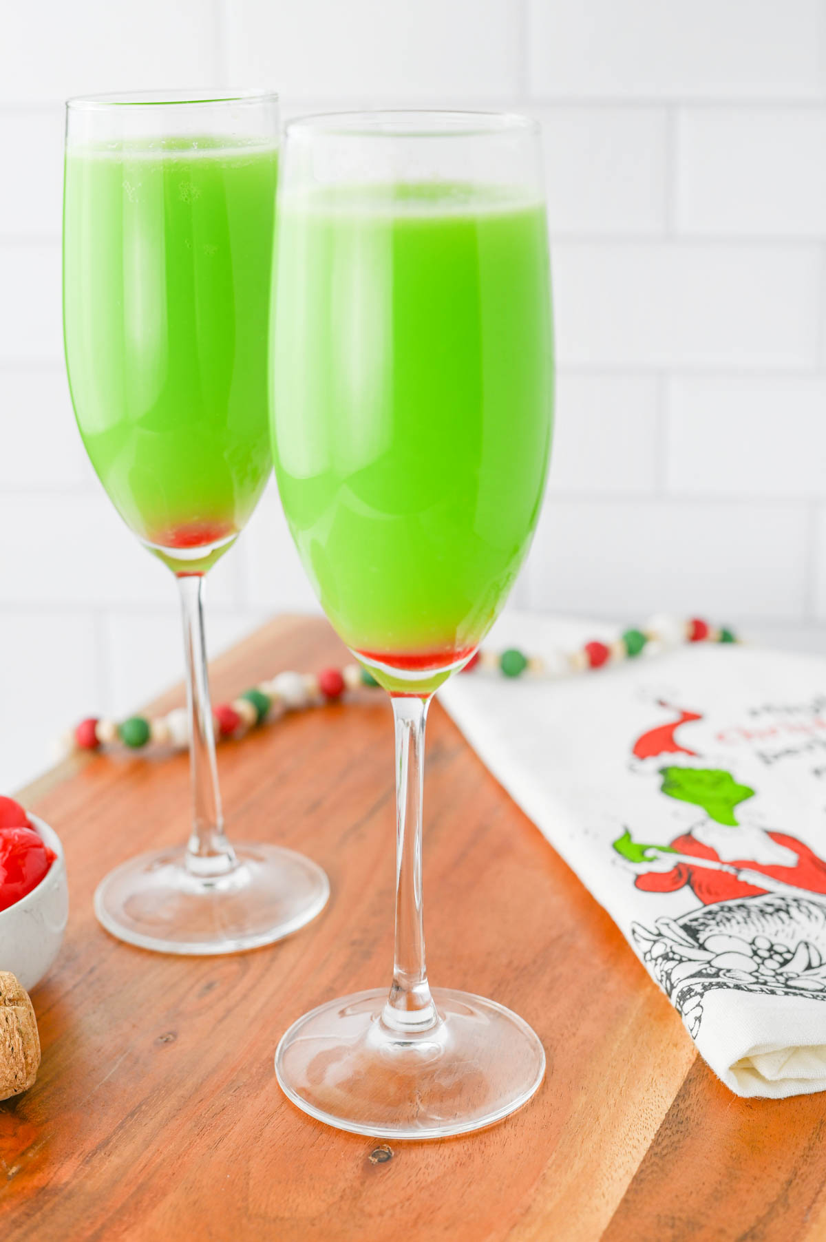 Green liquid in a champagne flute with red cherry in bottom of glass, with Grinch napkin on wood board.