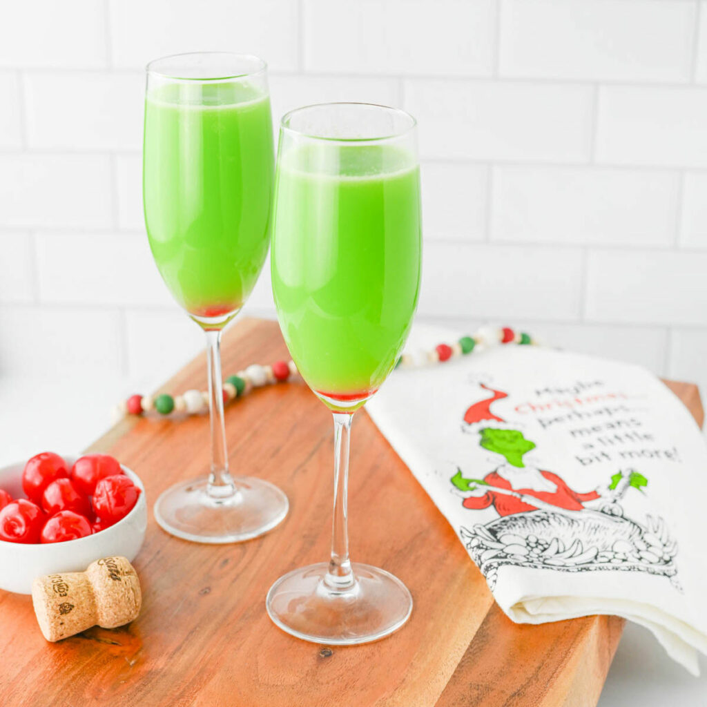 Green liquid in a champagne flute with red cherry in bottom of glass, with Grinch napkin on wood board.