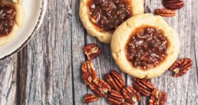 Two pecan pie cookies on a wood board with pecans scattered and a baking sheet of cookies in background.