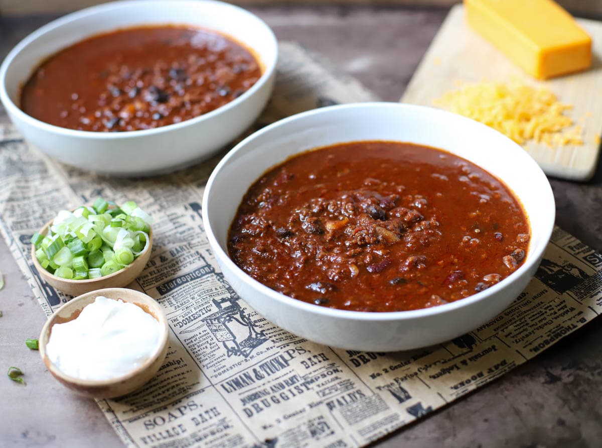 Two white bowls filled with chili on a newspaper with small bowls of green onions, sour cream, and a block  of cheese with shredded cheese.