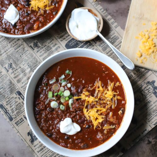 Two white bowls filled with chili and topped with spring onions, sour creme, and cheese on a newspaper with small bowls of green onions, sour cream, and a block of cheese with shredded cheese