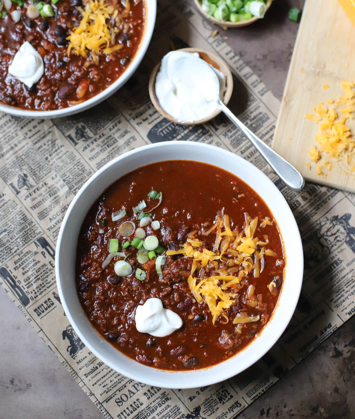 Two white bowls filled with chili and topped with spring onions, sour creme, and cheese on a newspaper with small bowls of green onions, sour cream, and a block  of cheese with shredded cheese