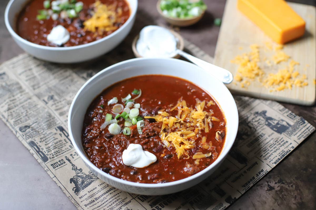 Two white bowls filled with chili and topped with spring onions, sour creme, and cheese on a newspaper with small bowls of green onions, sour cream, and a block  of cheese with shredded cheese.