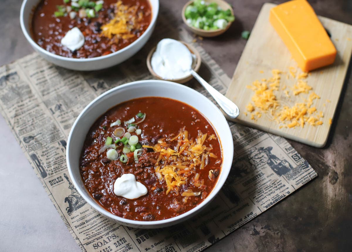 Two white bowls filled with chili and topped with spring onions, sour creme, and cheese on a newspaper with small bowls of green onions, sour cream, and a block  of cheese with shredded cheese