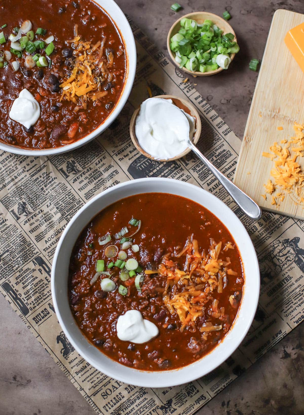 Two white bowls filled with chili and topped with spring onions, sour creme, and cheese on a newspaper with small bowls of green onions, sour cream, and a block  of cheese with shredded cheese.