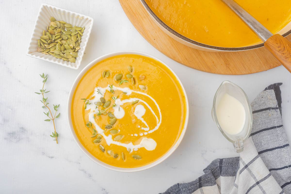 Pumpkin soup in white bowl with pumpkin seeds and thyme and coconut milk drizzled on top.