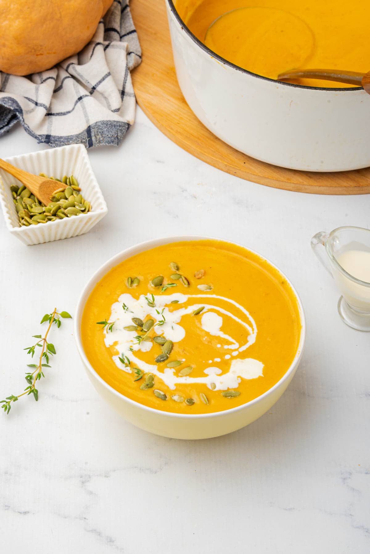 Pumpkin soup in white bowl on white counter with drizzle of coconut milk, pumpkin seeds, and thyme on top with bowl of pumpkin seeds, pot of soup, and a pumpkin in background.