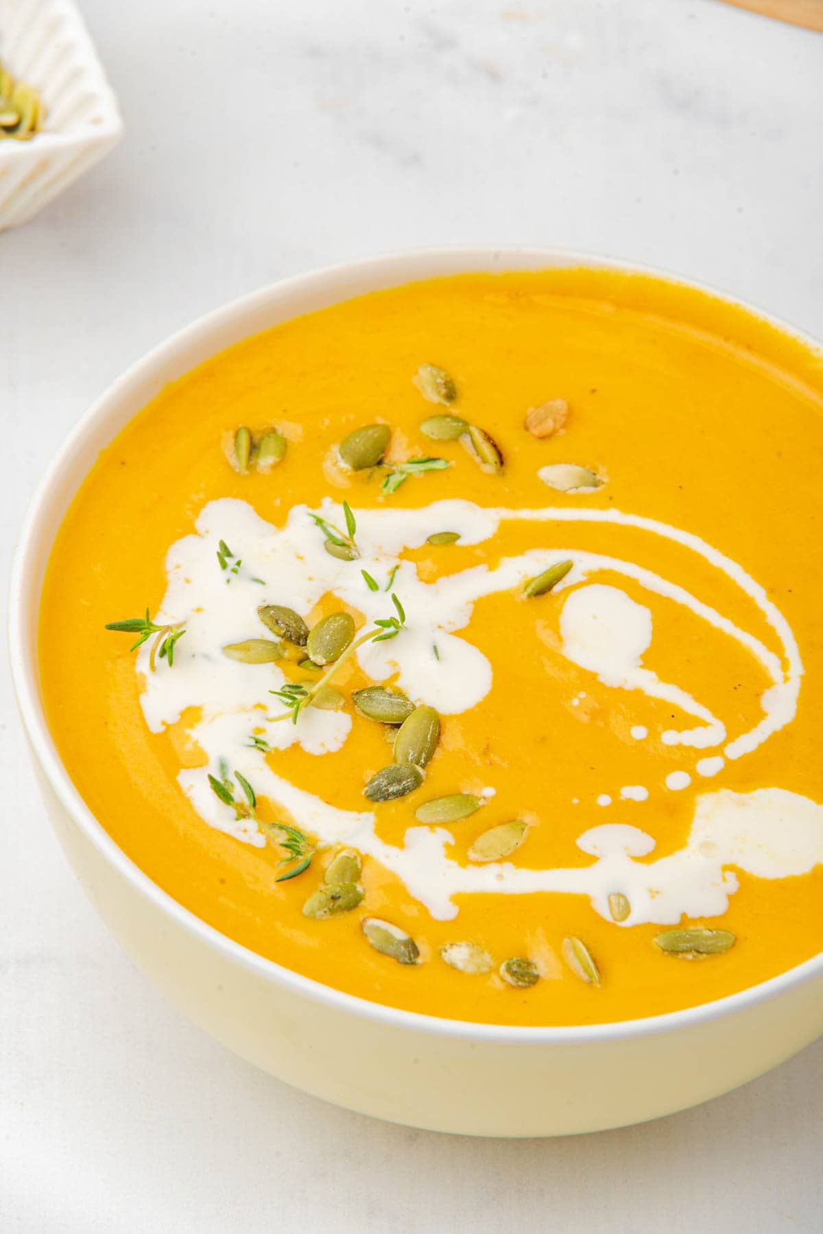 Pumpkin soup in white bowl on white counter with drizzle of coconut milk, pumpkin seeds, and thyme on top.