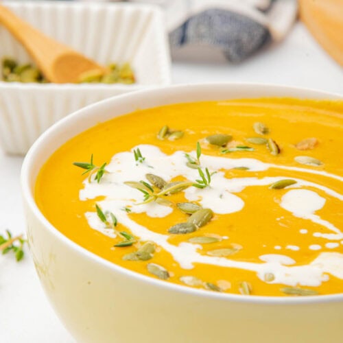 Pumpkin soup in white bowl on white counter with drizzle of coconut milk, pumpkin seeds, and thyme on top with bowl of pumpkin seeds, a kitchen towel, and a pumpkin in background.