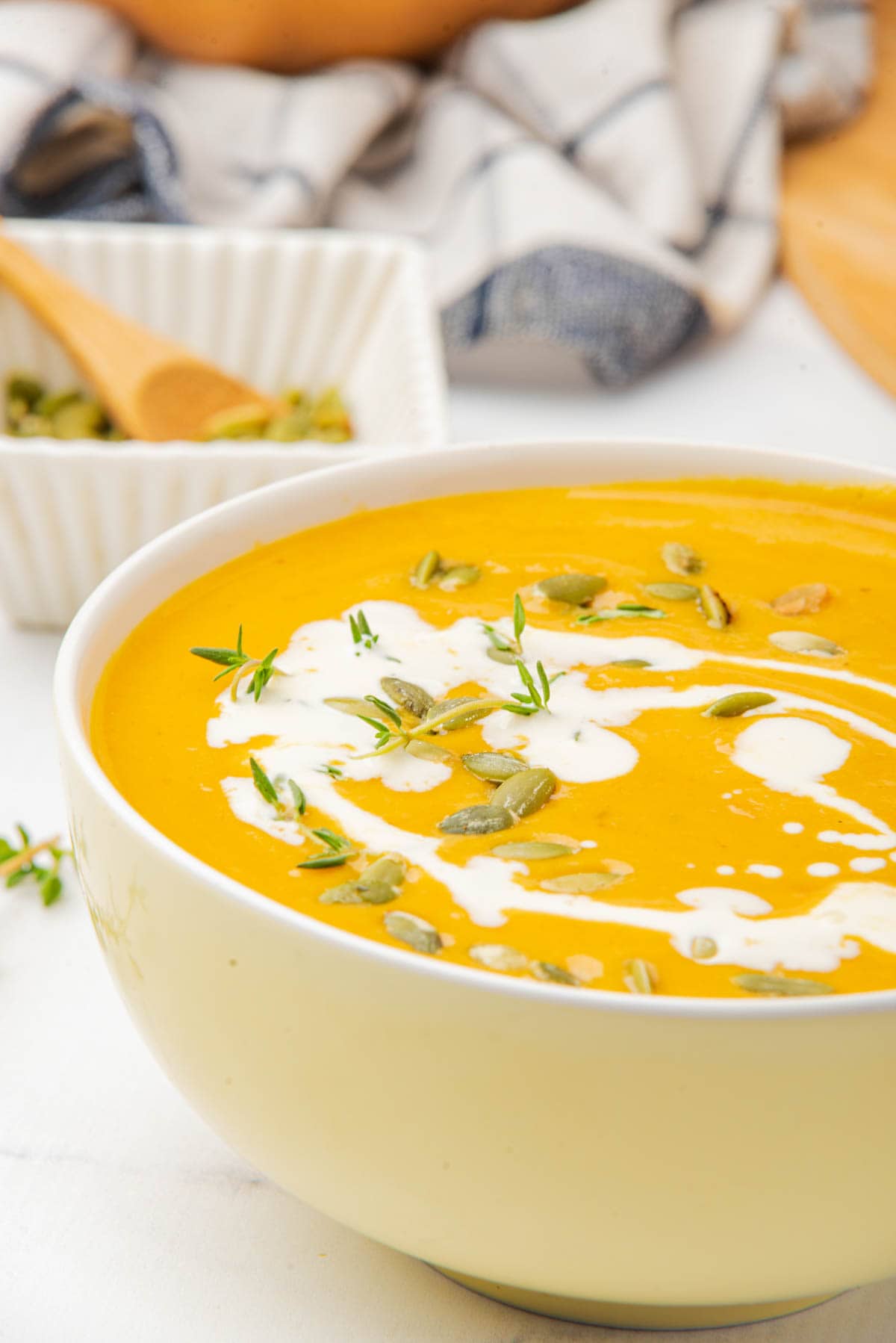 Pumpkin soup in white bowl on white counter with drizzle of coconut milk, pumpkin seeds, and thyme on top with bowl of pumpkin seeds, a kitchen towel, and a pumpkin in background.