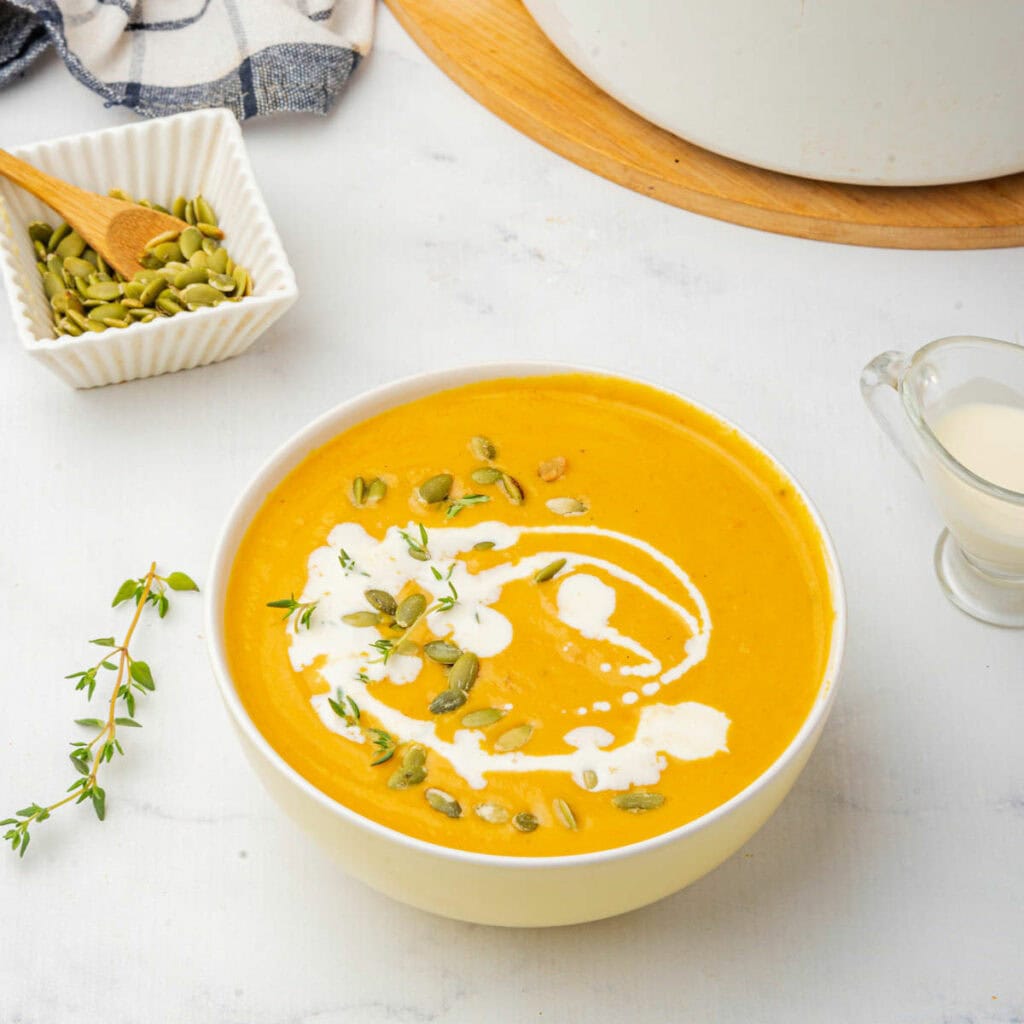 Pumpkin soup in white bowl on white counter with drizzle of coconut milk, pumpkin seeds, and thyme on top with bowl of pumpkin seeds, a kitchen towel, and a pot in background.