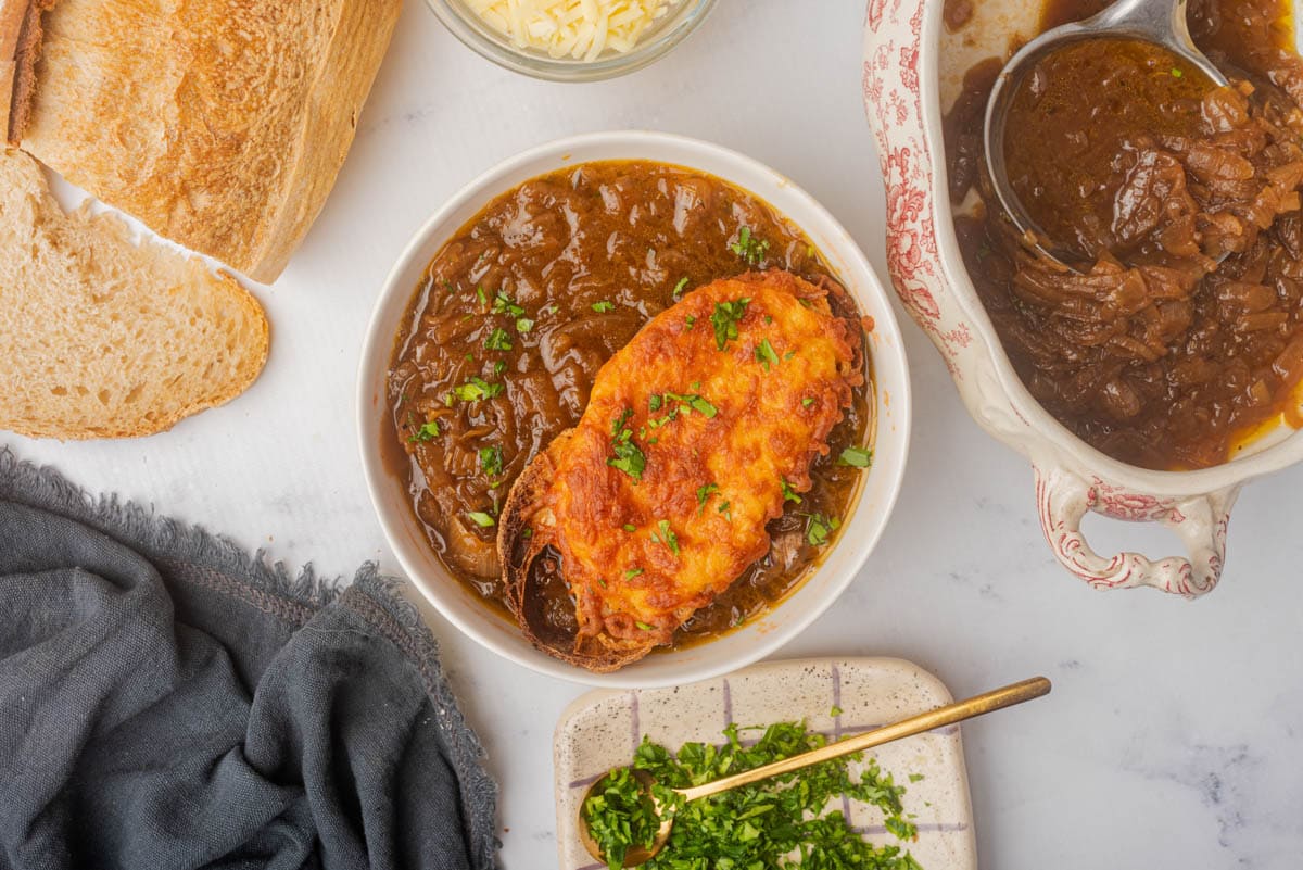 Bowl of French onion soup topped with cheese toast and sliced green onions, with bread to one side and green onions on plate with a spoon to the side.