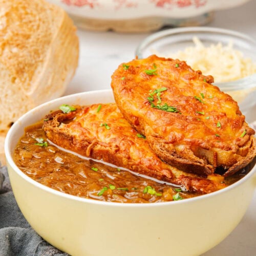 Bowl of French onion soup topped with cheese toast and sliced green onions, with bread to one side and green onions on plate with a spoon to the side.