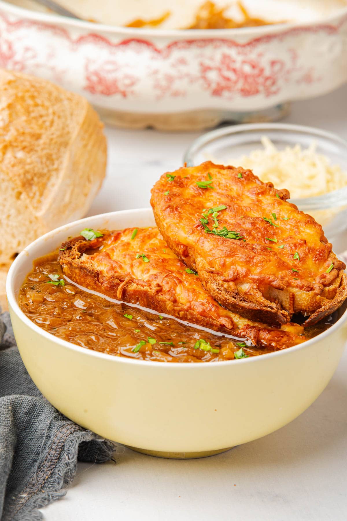 Bowl of French onion soup topped with cheese toast and sliced green onions, with bread to one side and green onions on plate with a spoon to the side.