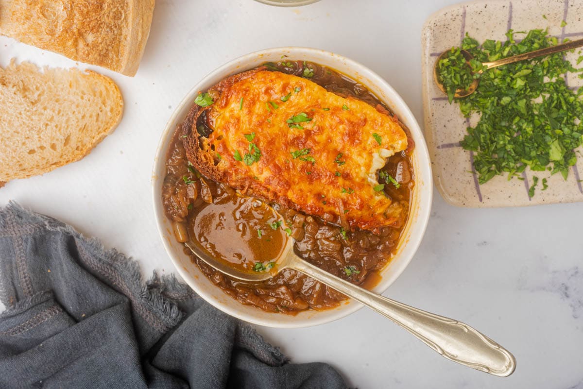 Bowl of French onion soup topped with cheese toast and sliced green onions, with bread to one side and green onions on plate with a spoon to the side.