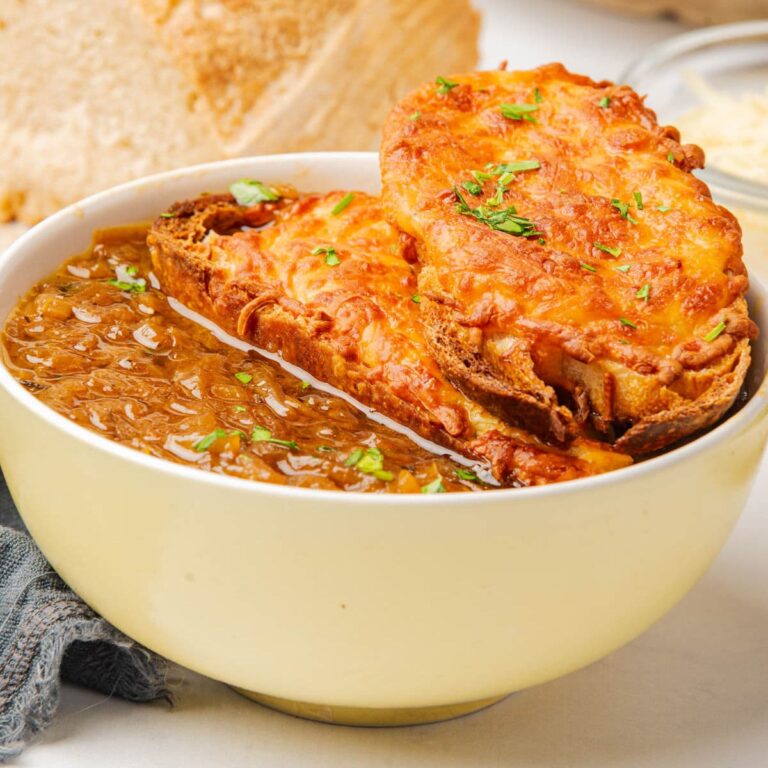 Bowl of French onion soup topped with cheese toast and sliced green onions, with bread to one side and green onions on plate with a spoon to the side.