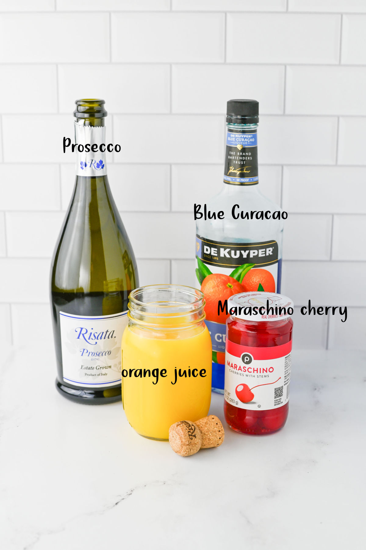 Bottles of Prosecco and Blue Curacao, jar of orange juice, and jar of cherries on a white counter.