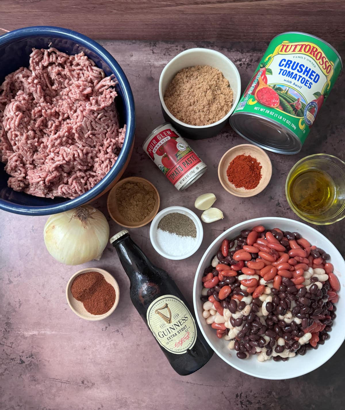 Ground beef, brown sugar, canned tomatoes, tomato paste, spices, beans, onion, garlic cloves, chili powder, salt and pepper, cumin, and olive oil on a brown counter.